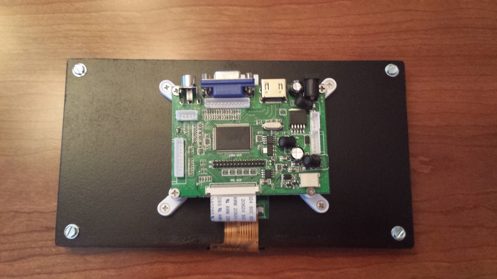 A piece of black quarter-inch MDF, with a circuit board attached using four plastic feet. A ribbon cable wraps around to the other side of the MDF, attaching to the screen on the backside.