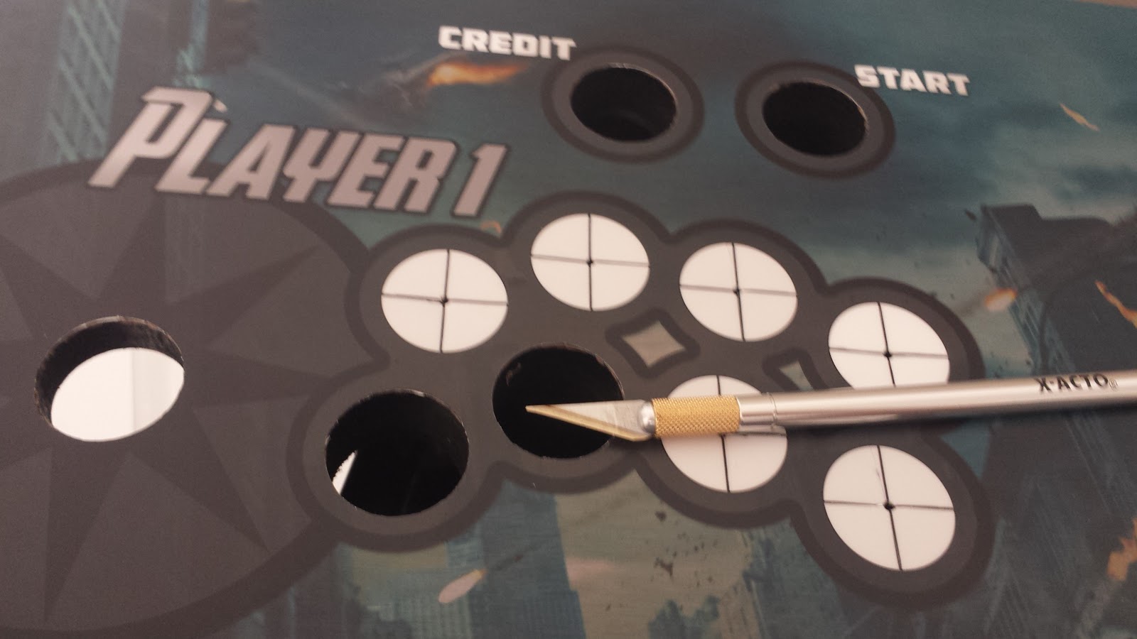 An X-acto Knife lays on top of the installed control panel artwork. Five of the 11 visible button & joystick holes have been cut away from the art, so the controls can be installed.